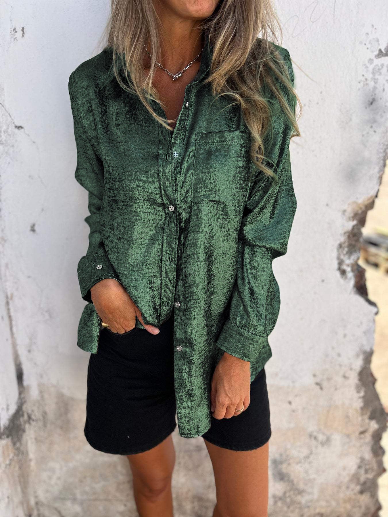 Woman wearing the Sophia Women's Shimmering Button-Up Blouse, a relaxed-fit blouse with a soft shimmer and long sleeves, styled with casual shorts and minimal jewelry, standing against a textured wall.