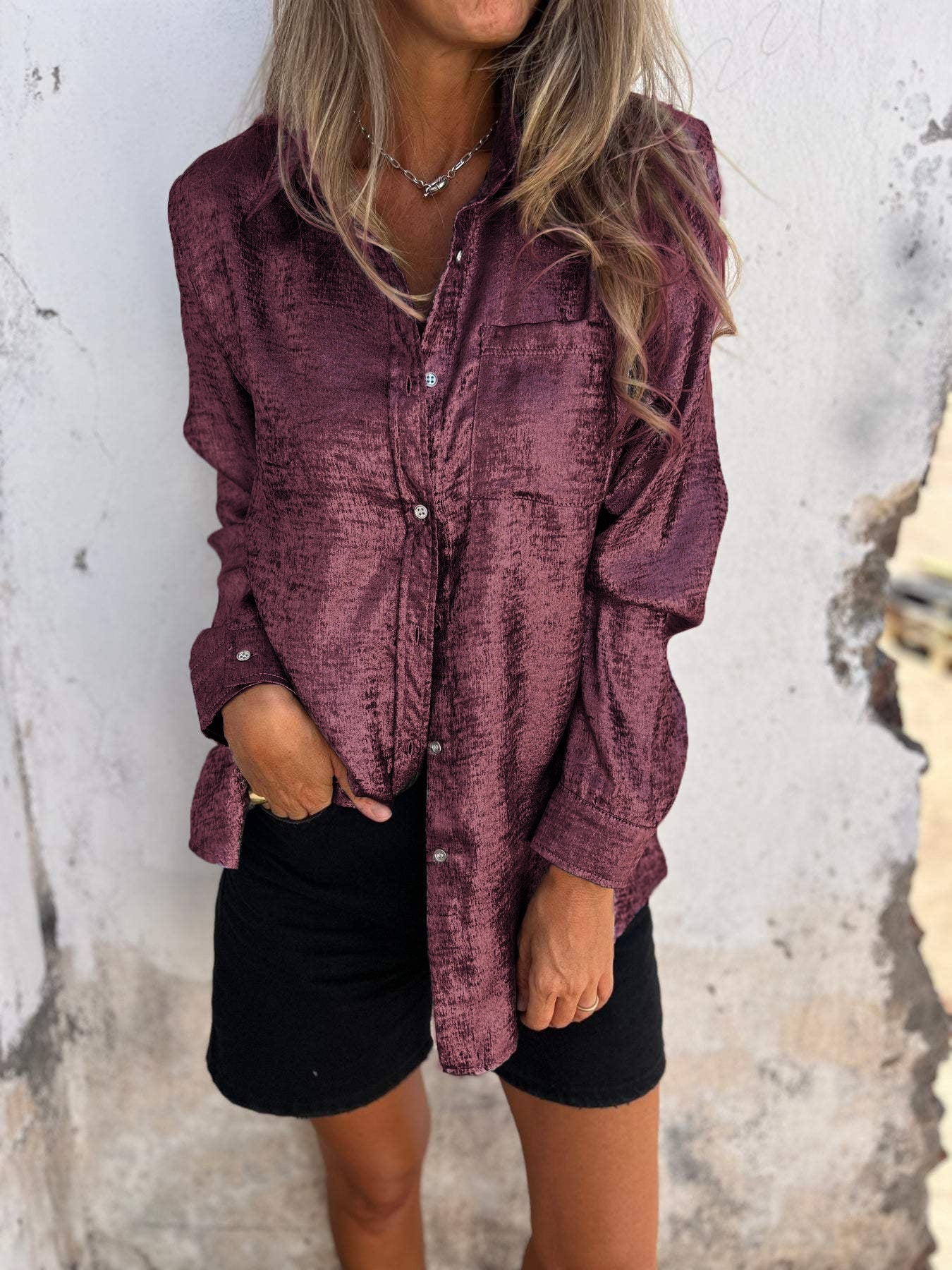 Woman wearing the Sophia Women's Shimmering Button-Up Blouse, a relaxed-fit blouse with a soft shimmer and long sleeves, styled with casual shorts and minimal jewelry, standing against a textured wall.