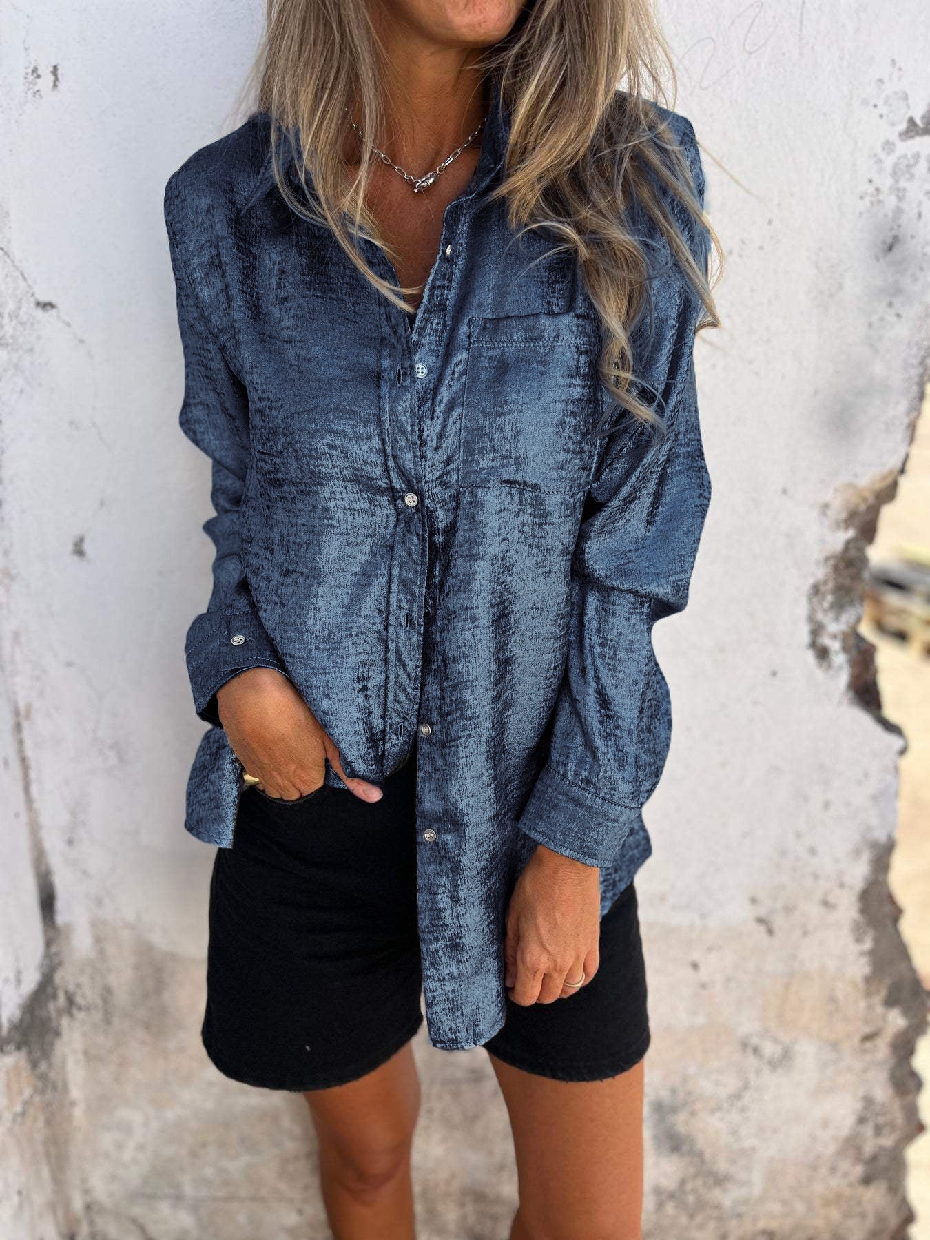 Woman wearing the Sophia Women's Shimmering Button-Up Blouse, a relaxed-fit blouse with a soft shimmer and long sleeves, styled with casual shorts and minimal jewelry, standing against a textured wall.