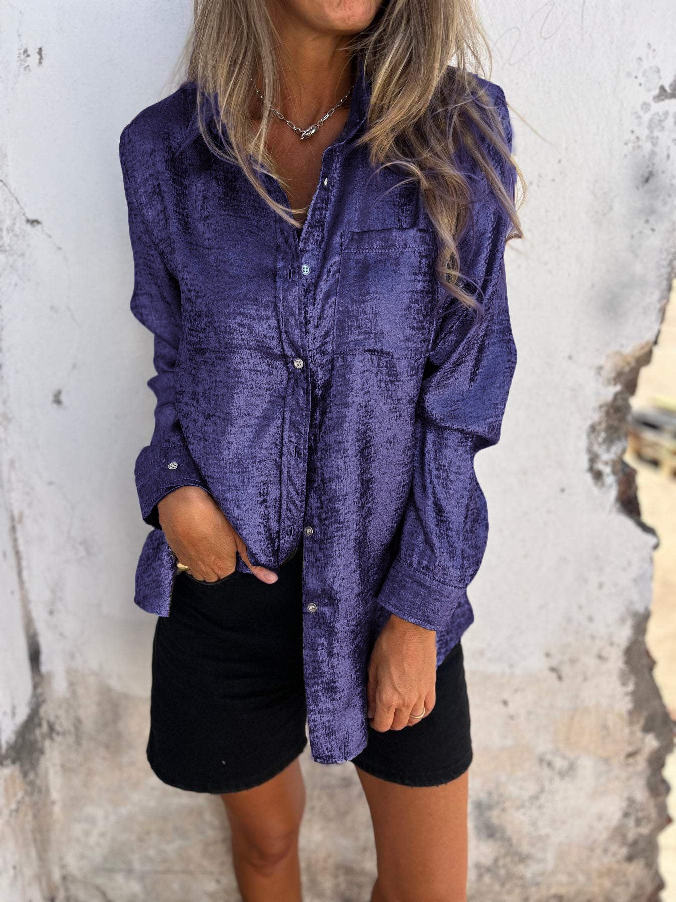 Woman wearing the Sophia Women's Shimmering Button-Up Blouse, a relaxed-fit blouse with a soft shimmer and long sleeves, styled with casual shorts and minimal jewelry, standing against a textured wall.