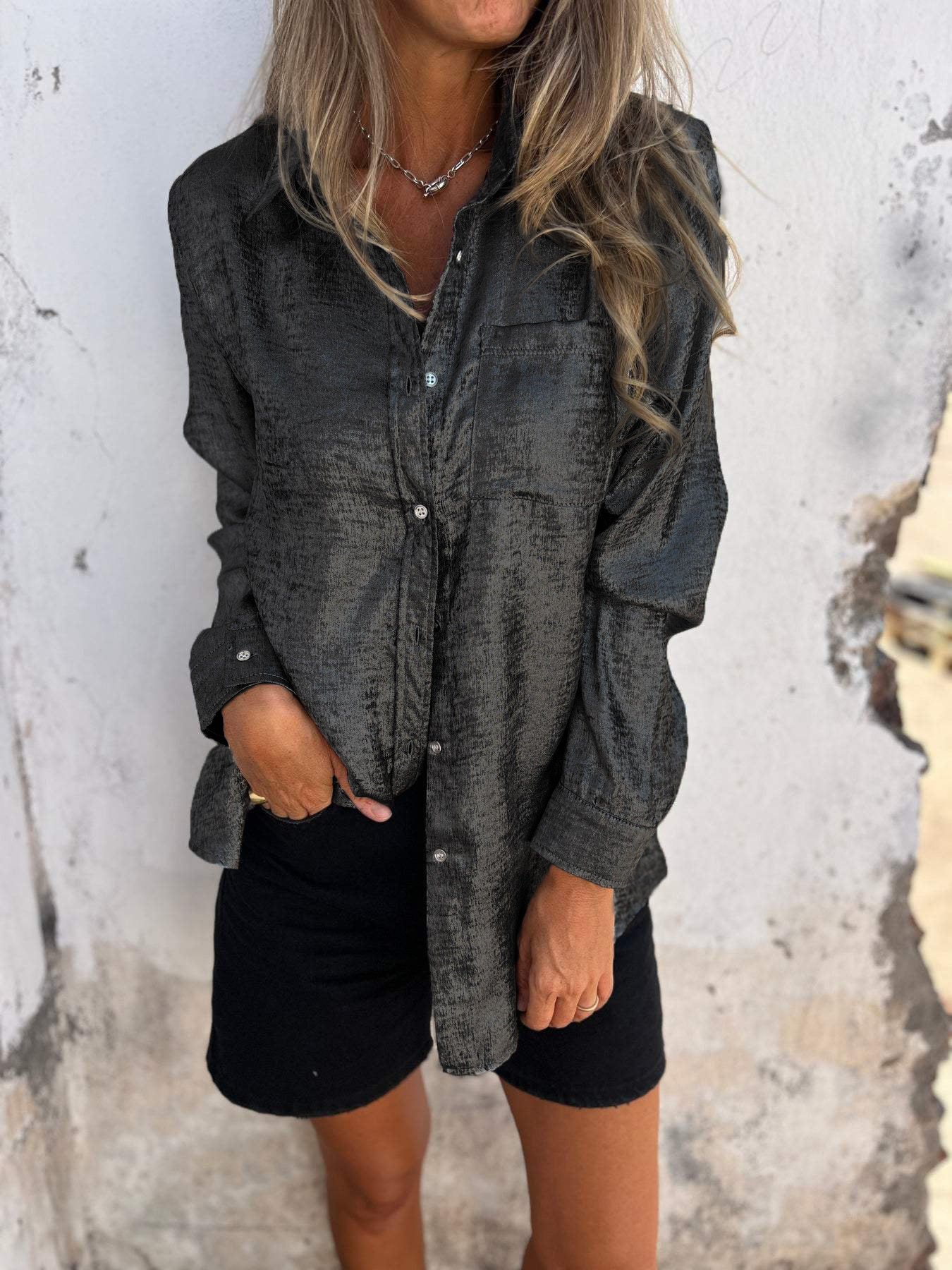 Woman wearing the Sophia Women's Shimmering Button-Up Blouse, a relaxed-fit blouse with a soft shimmer and long sleeves, styled with casual shorts and minimal jewelry, standing against a textured wall.