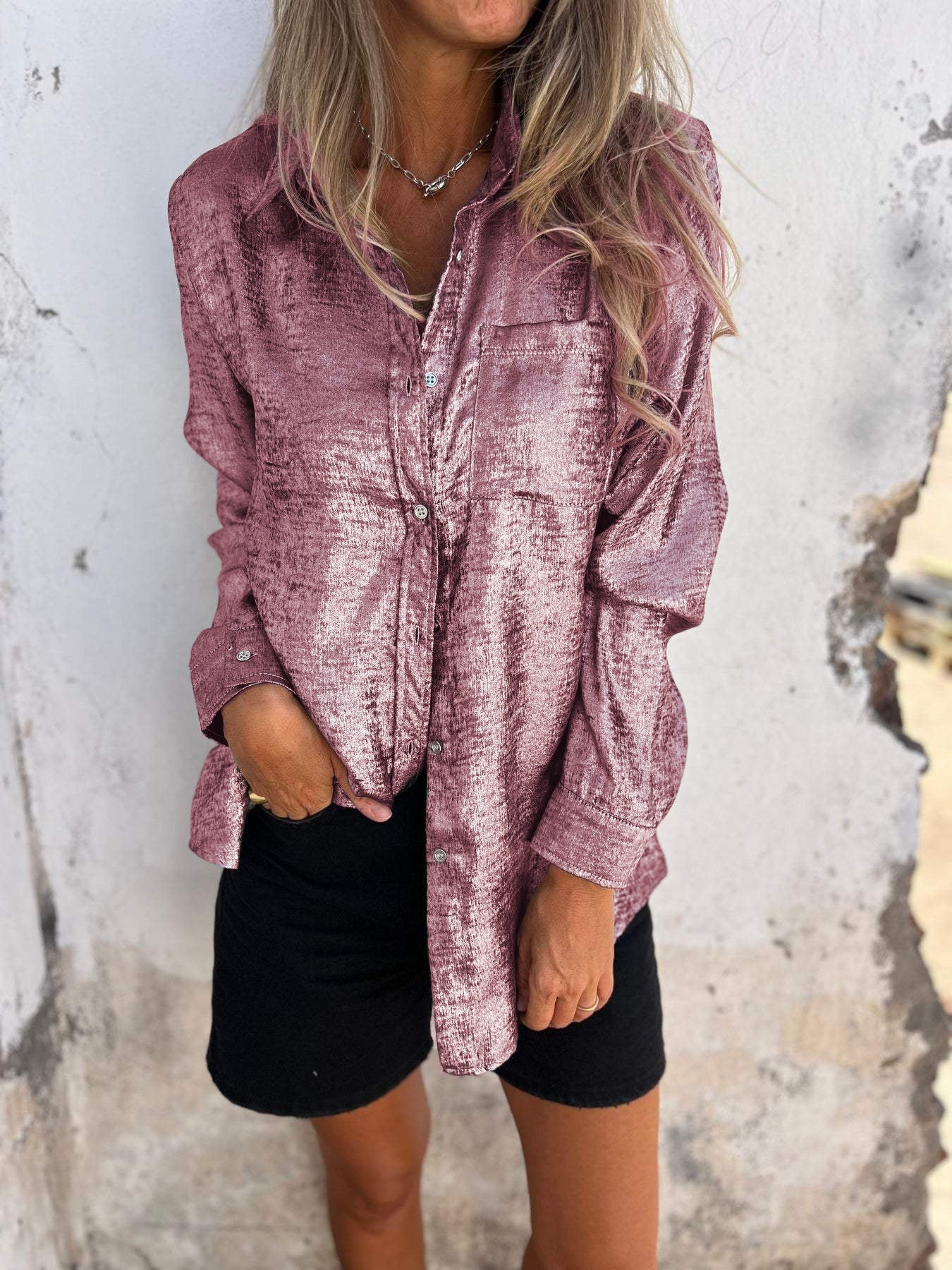 Woman wearing the Sophia Women's Shimmering Button-Up Blouse, a relaxed-fit blouse with a soft shimmer and long sleeves, styled with casual shorts and minimal jewelry, standing against a textured wall.