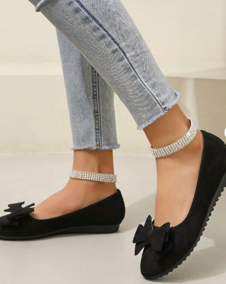 Elegant women's flats with a bow design, crafted for comfort and style, displayed on a textured surface with decorative floral accents nearby.