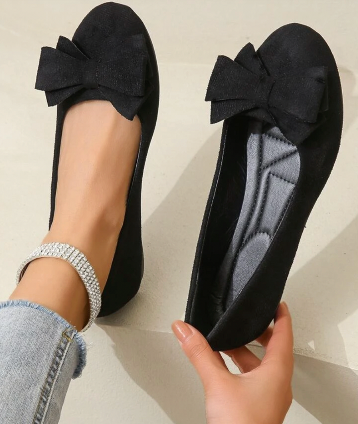 Elegant women's flats with a bow design, crafted for comfort and style, displayed on a textured surface with decorative floral accents nearby.