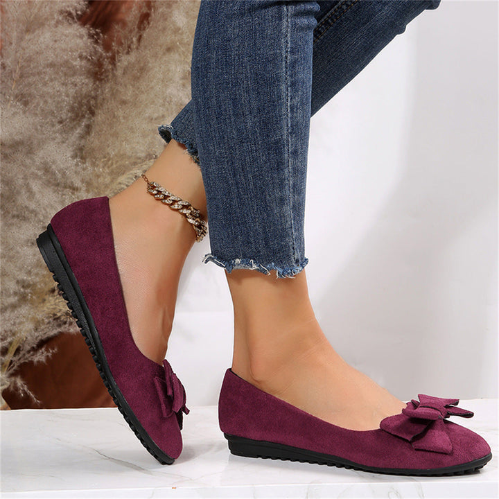 Elegant women's flats with a bow design, crafted for comfort and style, displayed on a textured surface with decorative floral accents nearby.