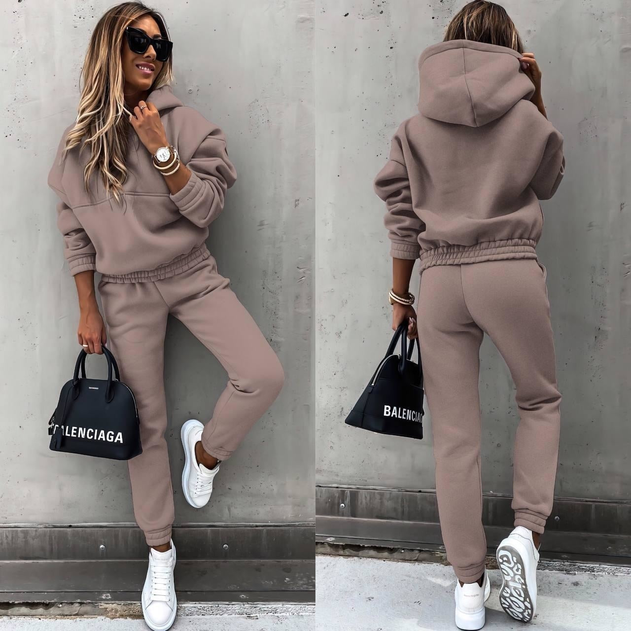 A woman wearing a casual two-piece tracksuit set with a hoodie and jogger-style pants. The outfit features a relaxed fit, perfect for both lounging and casual outings. She pairs it with white sneakers and a stylish handbag, creating a comfortable yet chic look.