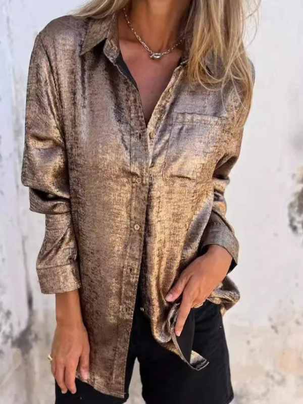 Woman wearing the Sophia Women's Shimmering Button-Up Blouse, a relaxed-fit blouse with a soft shimmer and long sleeves, styled with casual shorts and minimal jewelry, standing against a textured wall.