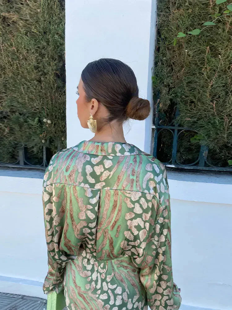 Woman wearing the Sofia Women's Satin Button-Up Dress, a luxurious dress with a bold, intricate print, styled with statement jewelry and a small textured handbag, posing elegantly outdoors against a lush green background.