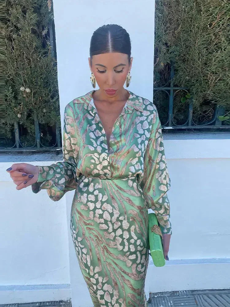 Woman wearing the Sofia Women's Satin Button-Up Dress, a luxurious dress with a bold, intricate print, styled with statement jewelry and a small textured handbag, posing elegantly outdoors against a lush green background.