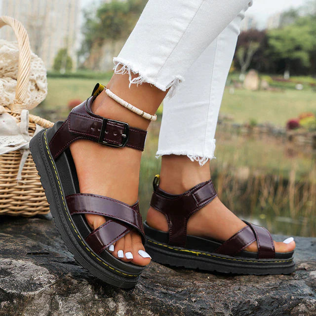 Close-up of women's strappy sandals with adjustable buckles, featuring a sturdy sole designed for outdoor activities, styled with white pants near a scenic background.