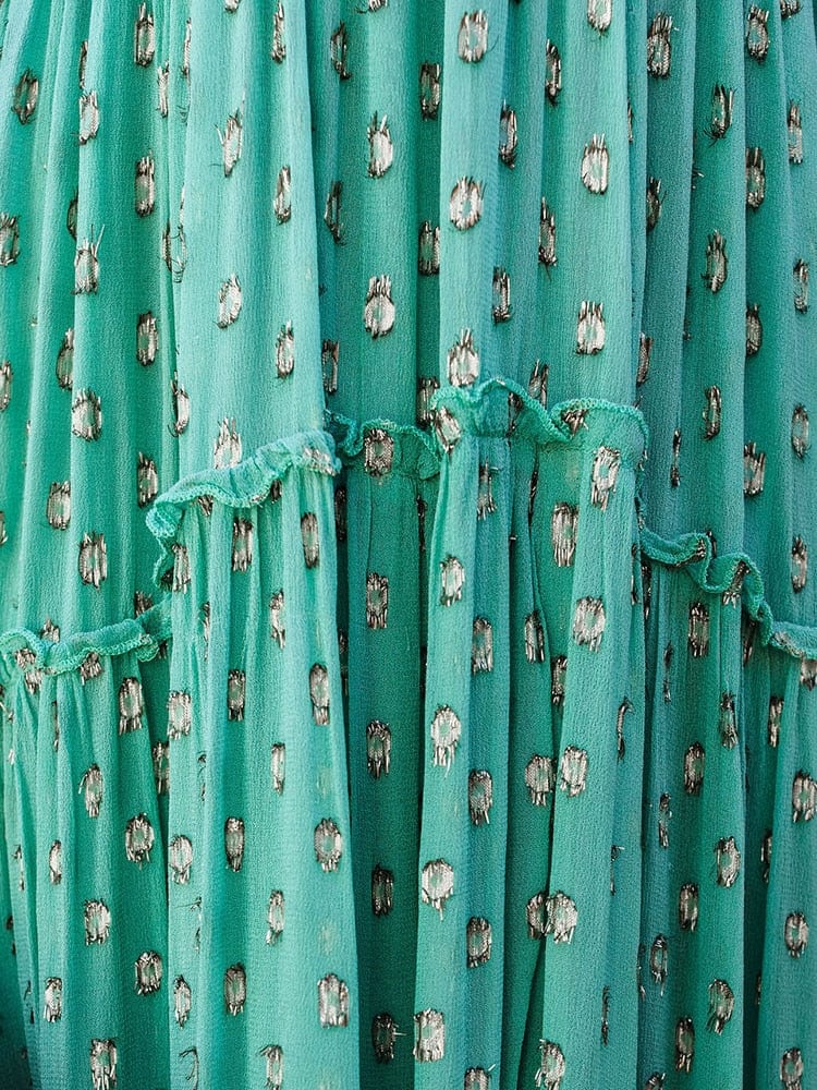 Woman wearing the Sienna Women's Tiered Maxi Dress, a long, flowy dress with adjustable straps and a bohemian-inspired tiered design, accessorized with layered necklaces and a clutch, posing gracefully against a textured wall with pampas grass in the background.