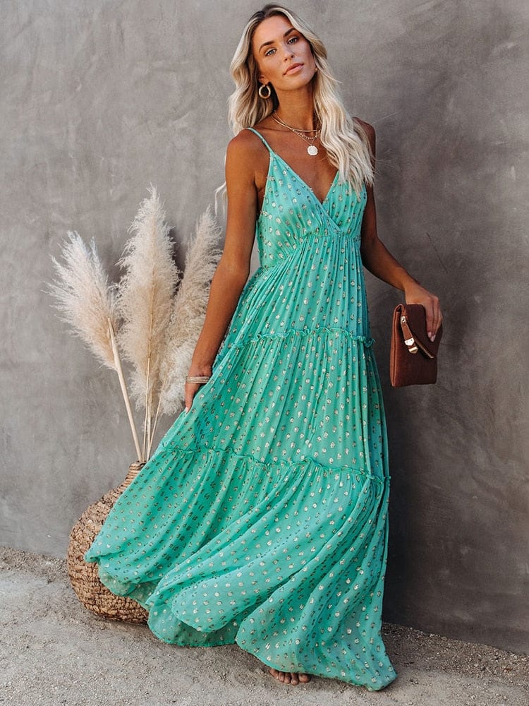 Woman wearing the Sienna Women's Tiered Maxi Dress, a long, flowy dress with adjustable straps and a bohemian-inspired tiered design, accessorized with layered necklaces and a clutch, posing gracefully against a textured wall with pampas grass in the background.