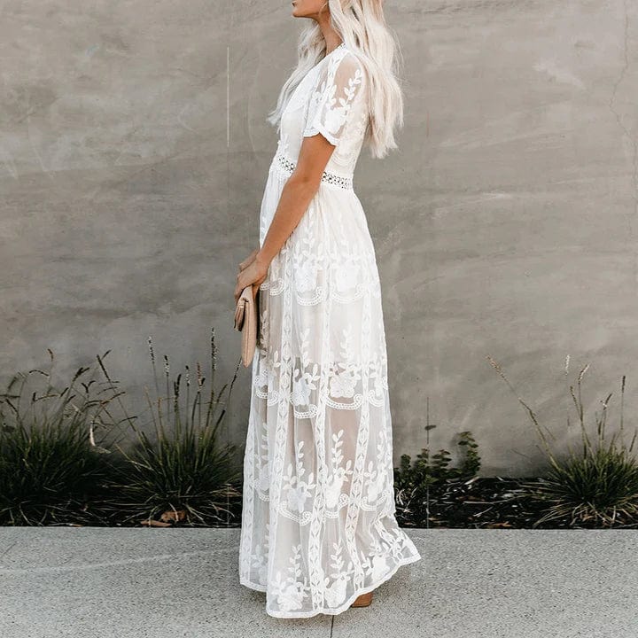 Woman wearing the Aurora Women's Lace Maxi Dress, a long dress with intricate lace detailing and a sheer, elegant design, standing against a textured wall with decorative vases nearby, exuding a graceful and sophisticated look.