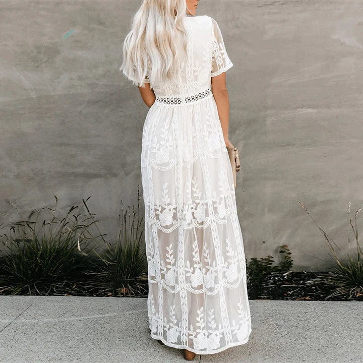 Woman wearing the Aurora Women's Lace Maxi Dress, a long dress with intricate lace detailing and a sheer, elegant design, standing against a textured wall with decorative vases nearby, exuding a graceful and sophisticated look.
