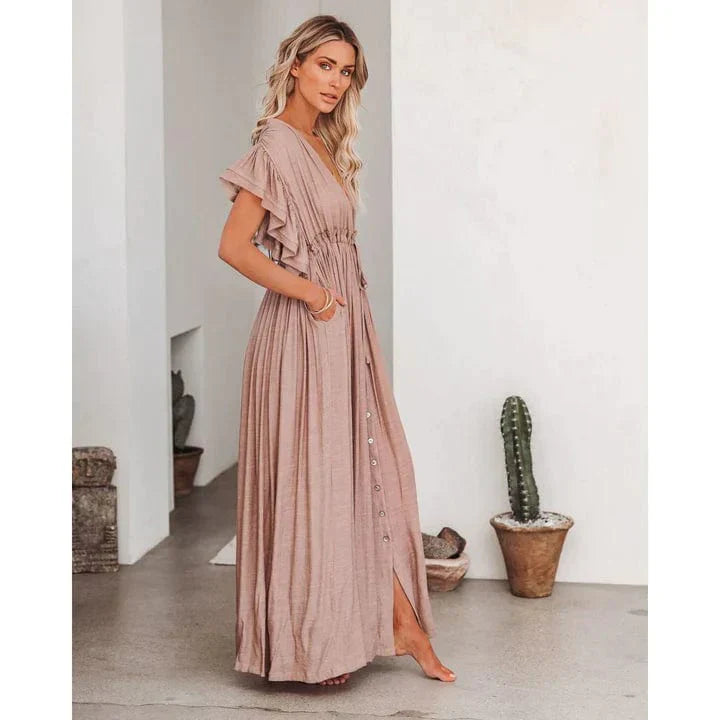 Woman wearing the Elena Women's Pleated Maxi Dress, a flowing dress with a graceful pleated design and ruffled sleeves, posing elegantly indoors against a minimalist background with a decorative cactus.