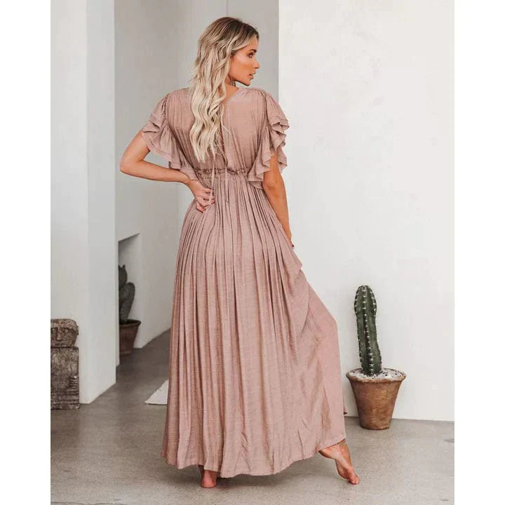 Woman wearing the Elena Women's Pleated Maxi Dress, a flowing dress with a graceful pleated design and ruffled sleeves, posing elegantly indoors against a minimalist background with a decorative cactus.