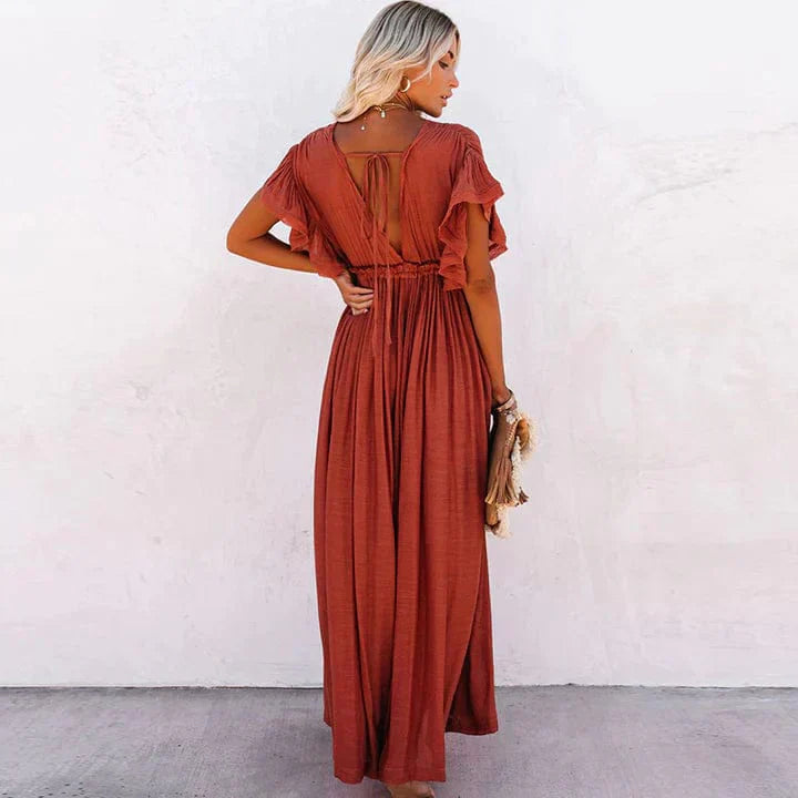 A woman sitting in a woven chair wearing a flowing boho maxi dress with a button-down front and cinched waist. The dress features a relaxed, loose fit and short sleeves, creating an effortlessly stylish and laid-back look. She accessorizes with layered necklaces and a chic handbag.