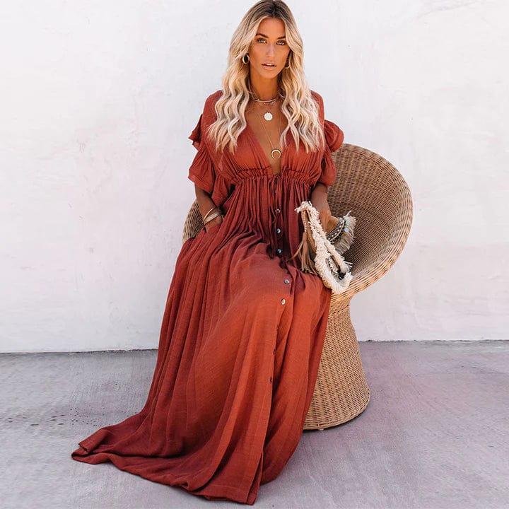 A woman sitting in a woven chair wearing a flowing boho maxi dress with a button-down front and cinched waist. The dress features a relaxed, loose fit and short sleeves, creating an effortlessly stylish and laid-back look. She accessorizes with layered necklaces and a chic handbag.