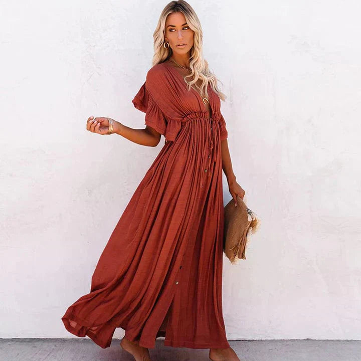 A woman sitting in a woven chair wearing a flowing boho maxi dress with a button-down front and cinched waist. The dress features a relaxed, loose fit and short sleeves, creating an effortlessly stylish and laid-back look. She accessorizes with layered necklaces and a chic handbag.