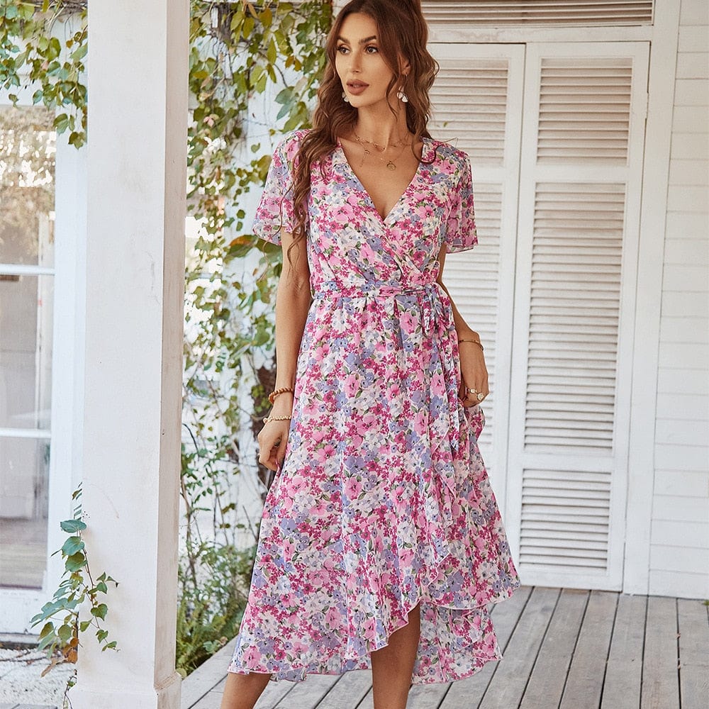 A woman wearing the Clara Women's Elegant Wrap Dress, a floral midi dress with a flattering wrap design, short sleeves, and an asymmetrical hemline, standing on a wooden porch surrounded by greenery.