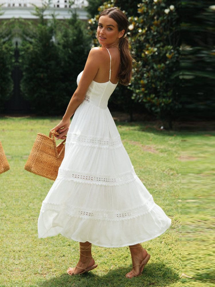 A woman walking in a garden wearing the Olivia | Strappy Tiered Midi Dress, featuring a flowy, tiered design with delicate straps. She holds a woven handbag, creating a chic, summery look perfect for outdoor occasions.