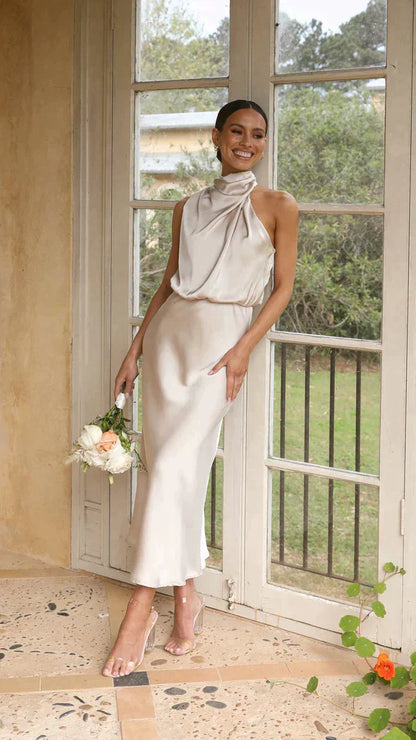 A woman wearing a sleeveless midi dress with an elegant draped neckline, standing outdoors in a stylish setting. The dress has a sleek and fitted silhouette, perfect for formal occasions or chic events. She is accessorized with minimalist heels for a polished, sophisticated look.