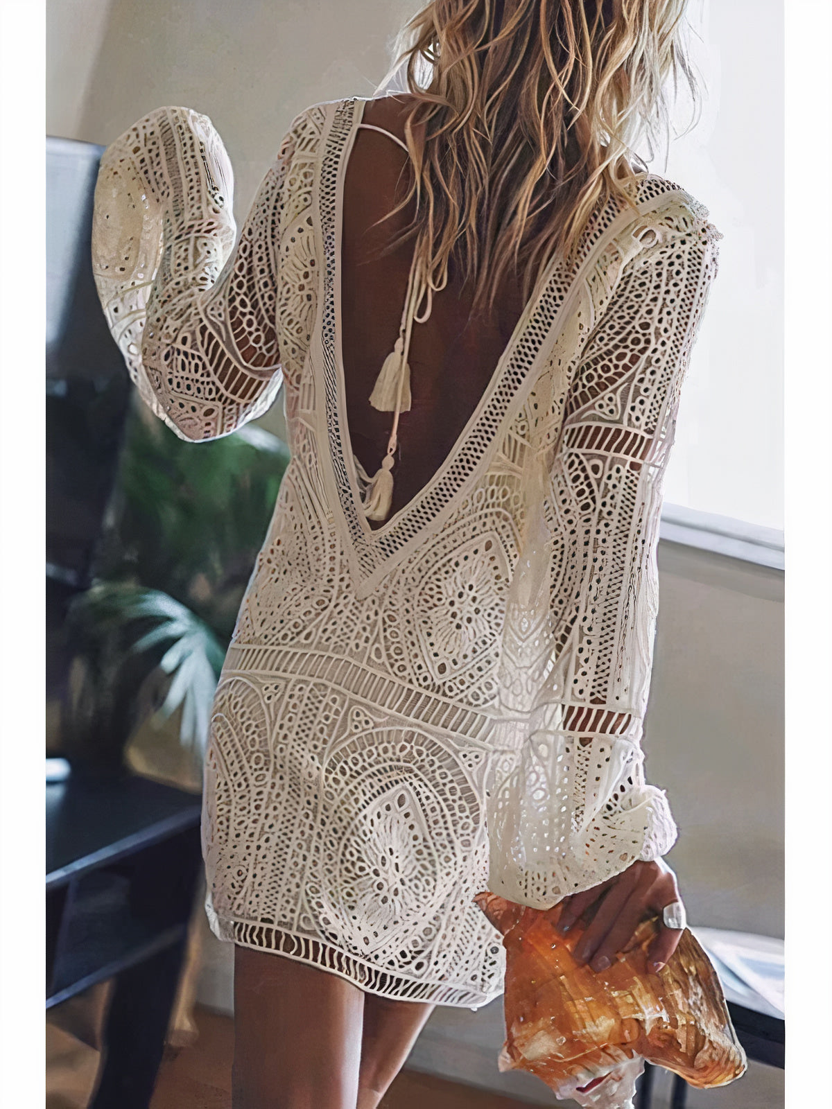 Rear view of a woman in an open-back lace dress with intricate bohemian detailing, holding a large seashell. The dress features a delicate tassel tie at the back and long sleeves, creating an effortless, beach-inspired look.