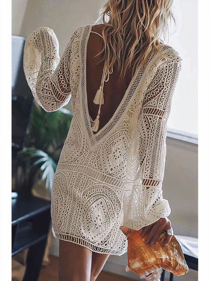 Rear view of a woman in an open-back lace dress with intricate bohemian detailing, holding a large seashell. The dress features a delicate tassel tie at the back and long sleeves, creating an effortless, beach-inspired look.