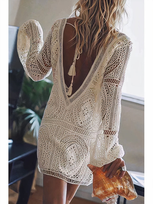 Rear view of a woman in an open-back lace dress with intricate bohemian detailing, holding a large seashell. The dress features a delicate tassel tie at the back and long sleeves, creating an effortless, beach-inspired look.