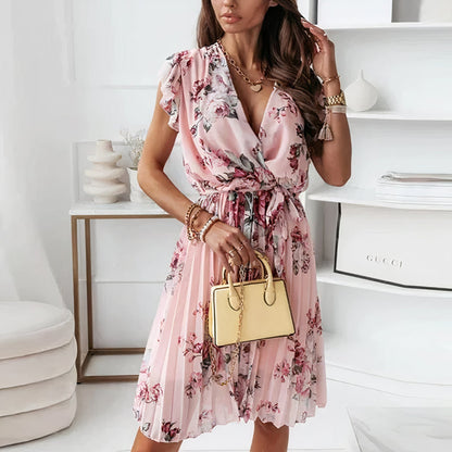 A woman wearing a floral wrap dress with ruffle sleeves, accessorized with a clutch and layered jewelry. The dress features a tied waist and pleated skirt, creating an elegant, feminine look ideal for casual gatherings or special occasions in a stylish, sophisticated setting.