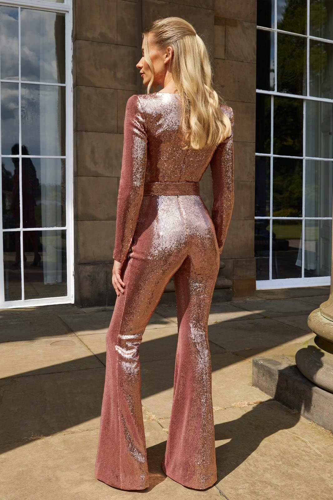 Woman wearing the Serena Women's Sequin Jumpsuit, a glamorous wide-leg jumpsuit with a shimmering sequin design, posing elegantly on a terrace at night with architectural buildings in the background.