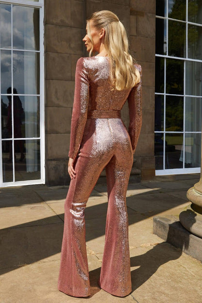 Woman wearing the Serena Women's Sequin Jumpsuit, a glamorous wide-leg jumpsuit with a shimmering sequin design, posing elegantly on a terrace at night with architectural buildings in the background.