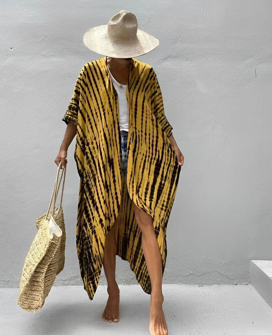 Woman wearing the Isla Women's Boho Kimono Cover-Up, a flowy kimono-style beachwear piece, styled over a swimsuit. She accessorizes with a wide-brimmed hat, a large woven tote bag, and sandals, creating a chic, relaxed beach look against a textured wall backdrop.