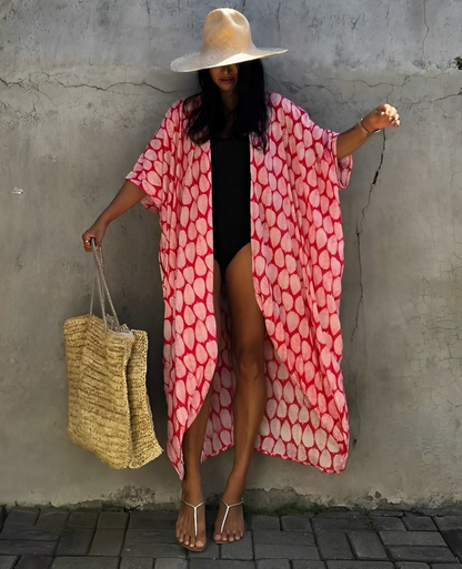 Woman wearing the Isla Women's Boho Kimono Cover-Up, a flowy kimono-style beachwear piece, styled over a swimsuit. She accessorizes with a wide-brimmed hat, a large woven tote bag, and sandals, creating a chic, relaxed beach look against a textured wall backdrop.