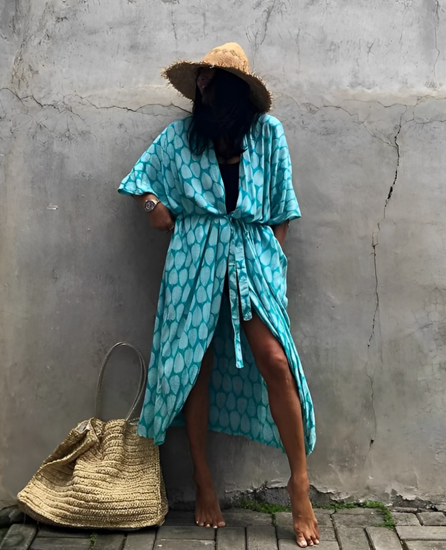 Woman wearing the Isla Women's Boho Kimono Cover-Up, a flowy kimono-style beachwear piece, styled over a swimsuit. She accessorizes with a wide-brimmed hat, a large woven tote bag, and sandals, creating a chic, relaxed beach look against a textured wall backdrop.