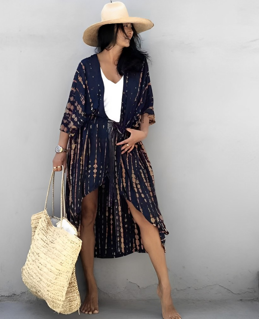 Woman wearing the Isla Women's Boho Kimono Cover-Up, a flowy kimono-style beachwear piece, styled over a swimsuit. She accessorizes with a wide-brimmed hat, a large woven tote bag, and sandals, creating a chic, relaxed beach look against a textured wall backdrop.