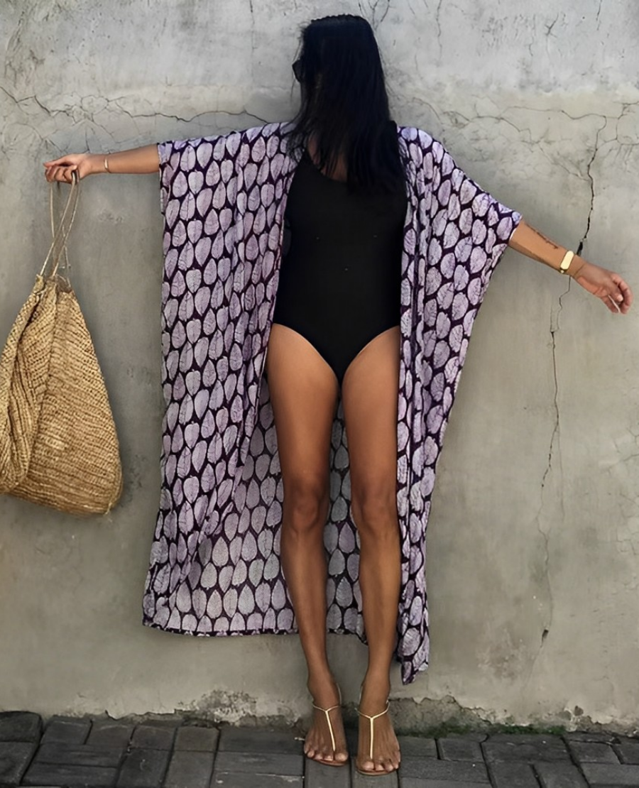 Woman wearing the Isla Women's Boho Kimono Cover-Up, a flowy kimono-style beachwear piece, styled over a swimsuit. She accessorizes with a wide-brimmed hat, a large woven tote bag, and sandals, creating a chic, relaxed beach look against a textured wall backdrop.