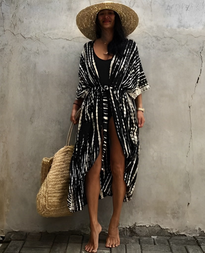 Woman wearing the Isla Women's Boho Kimono Cover-Up, a flowy kimono-style beachwear piece, styled over a swimsuit. She accessorizes with a wide-brimmed hat, a large woven tote bag, and sandals, creating a chic, relaxed beach look against a textured wall backdrop.