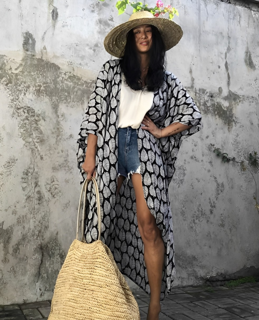 Woman wearing the Isla Women's Boho Kimono Cover-Up, a flowy kimono-style beachwear piece, styled over a swimsuit. She accessorizes with a wide-brimmed hat, a large woven tote bag, and sandals, creating a chic, relaxed beach look against a textured wall backdrop.