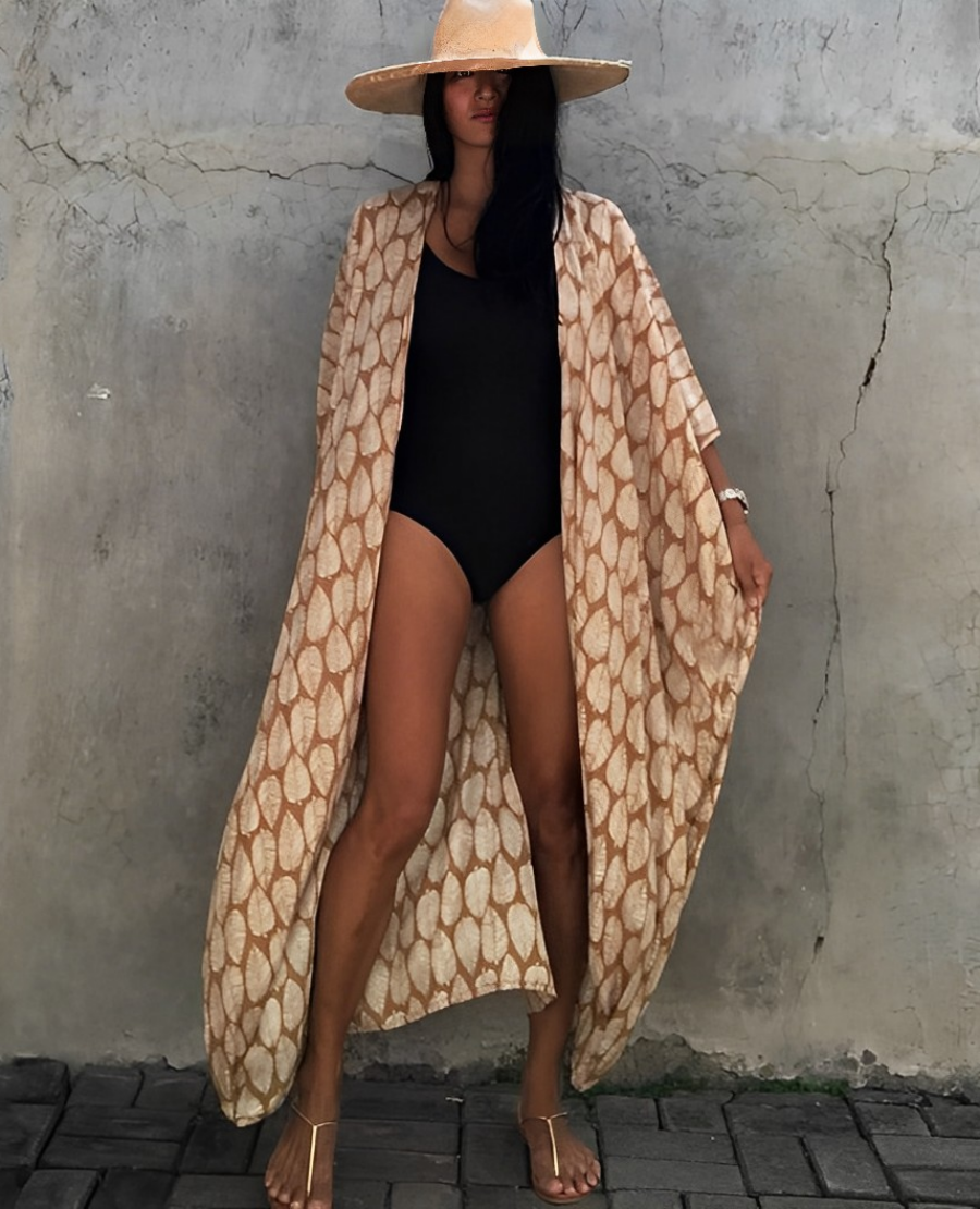 Woman wearing the Isla Women's Boho Kimono Cover-Up, a flowy kimono-style beachwear piece, styled over a swimsuit. She accessorizes with a wide-brimmed hat, a large woven tote bag, and sandals, creating a chic, relaxed beach look against a textured wall backdrop.