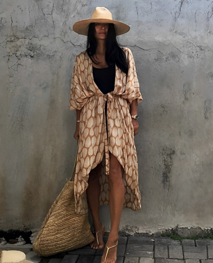 Woman wearing the Isla Women's Boho Kimono Cover-Up, a flowy kimono-style beachwear piece, styled over a swimsuit. She accessorizes with a wide-brimmed hat, a large woven tote bag, and sandals, creating a chic, relaxed beach look against a textured wall backdrop.