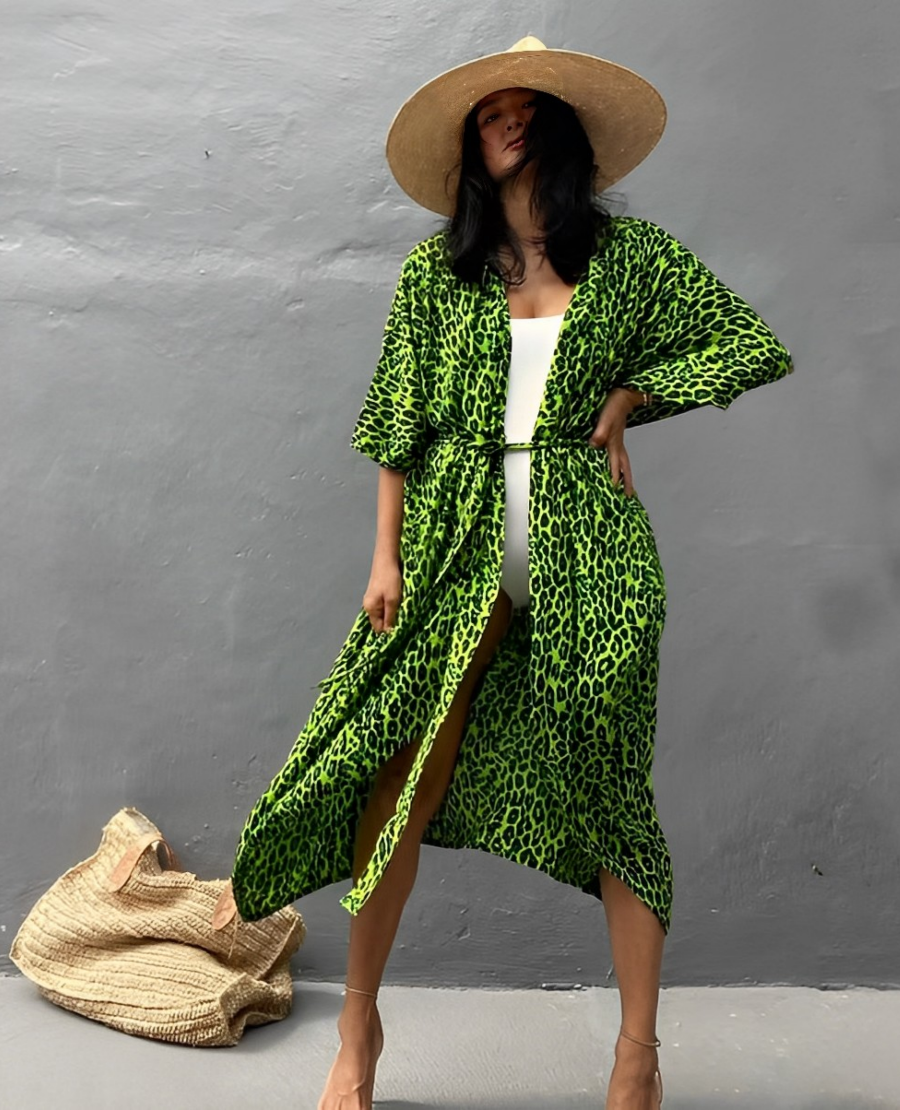 Woman wearing the Isla Women's Boho Kimono Cover-Up, a flowy kimono-style beachwear piece, styled over a swimsuit. She accessorizes with a wide-brimmed hat, a large woven tote bag, and sandals, creating a chic, relaxed beach look against a textured wall backdrop.