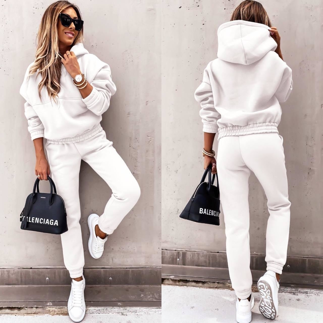 A woman wearing a casual two-piece tracksuit set with a hoodie and jogger-style pants. The outfit features a relaxed fit, perfect for both lounging and casual outings. She pairs it with white sneakers and a stylish handbag, creating a comfortable yet chic look.