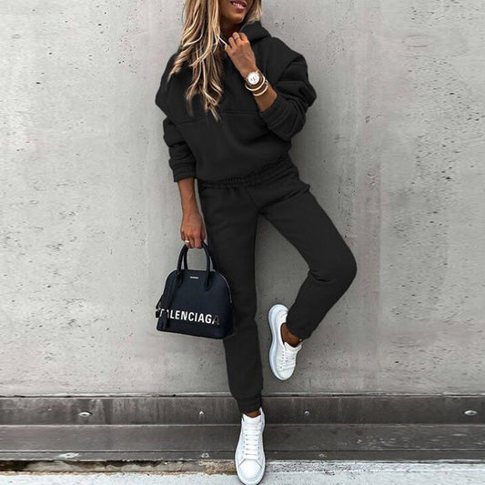 A woman wearing a casual two-piece tracksuit set with a hoodie and jogger-style pants. The outfit features a relaxed fit, perfect for both lounging and casual outings. She pairs it with white sneakers and a stylish handbag, creating a comfortable yet chic look.