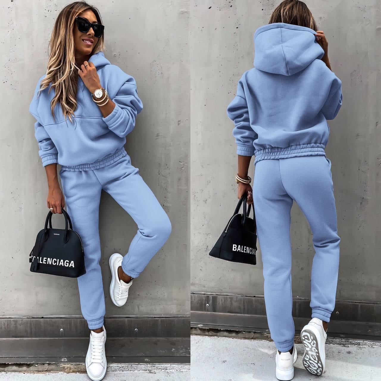 A woman wearing a casual two-piece tracksuit set with a hoodie and jogger-style pants. The outfit features a relaxed fit, perfect for both lounging and casual outings. She pairs it with white sneakers and a stylish handbag, creating a comfortable yet chic look.