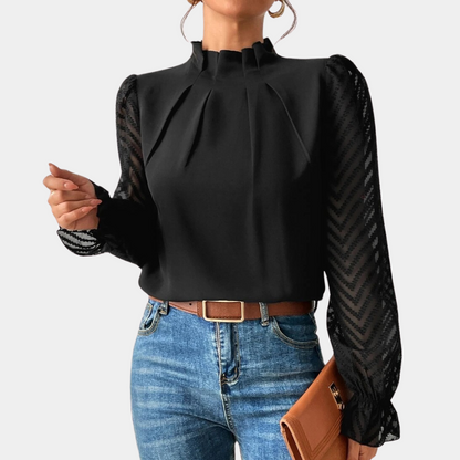 Woman wearing an elegant blouse with a high neckline, pleated front detail, sheer long sleeves with a subtle pattern, and ruffled cuffs, paired with denim jeans for a stylish and sophisticated look.