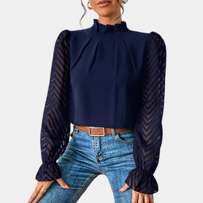 Woman wearing an elegant blouse with a high neckline, pleated front detail, sheer long sleeves with a subtle pattern, and ruffled cuffs, paired with denim jeans for a stylish and sophisticated look.