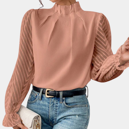 Woman wearing an elegant blouse with a high neckline, pleated front detail, sheer long sleeves with a subtle pattern, and ruffled cuffs, paired with denim jeans for a stylish and sophisticated look.