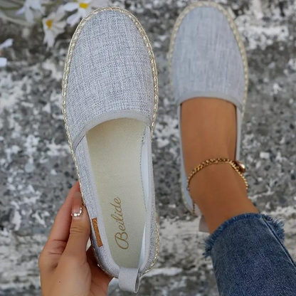 Close-up of a woman's hand holding a pair of slip-on shoes with a textured fabric design. One shoe is on her foot, showcasing a comfortable and casual style ideal for everyday wear.