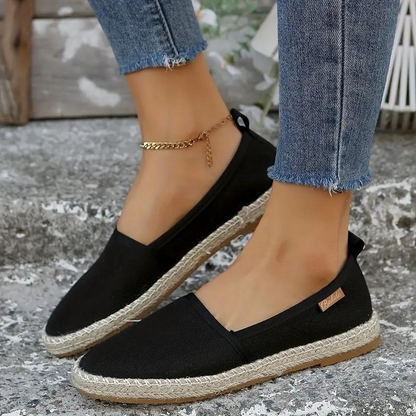 Close-up of a woman's hand holding a pair of slip-on shoes with a textured fabric design. One shoe is on her foot, showcasing a comfortable and casual style ideal for everyday wear.